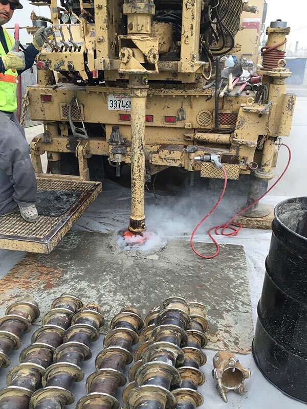 A drill rig advancing a boring through a concrete surface, with rows of soil sampling sleeves laid out in the foreground and a field technician at the controls.